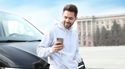 A man using his phone leaning on the hood of his vehicle.